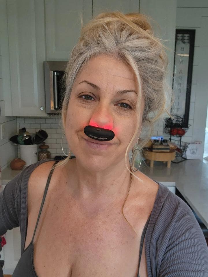 Woman using NoMore® Colds red light therapy device in her nose at home, Michelle Bloom review saying she stopped Advil after one month, stayed healthy even when her husband got sick, and had great results